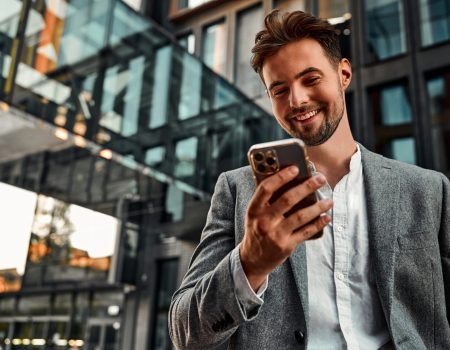 Portrait of attractive confident modern smiling happy adult dressed gray suit man holding a phone and standing outside near office building. Online work, communication. Copy space.